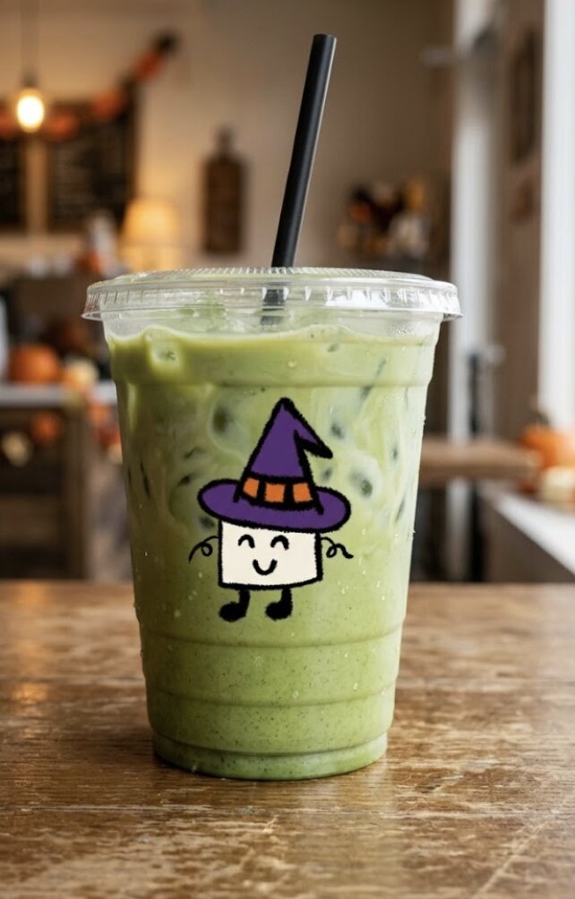 Version générée avec le prompt anglais : la mascotte sorcière, plus petite, imprimée sur un gobelet de matcha glacé posé sur une table en bois dans un coffee shop flou